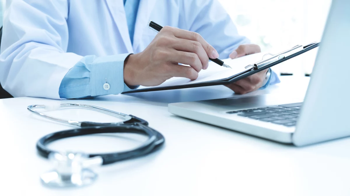Médico preenchendo formulário em prancheta ao lado de estetoscópio e notebook, representando análise de documentos para Isenção de IR e cuidados para evitar que o pedido de isenção seja negado.