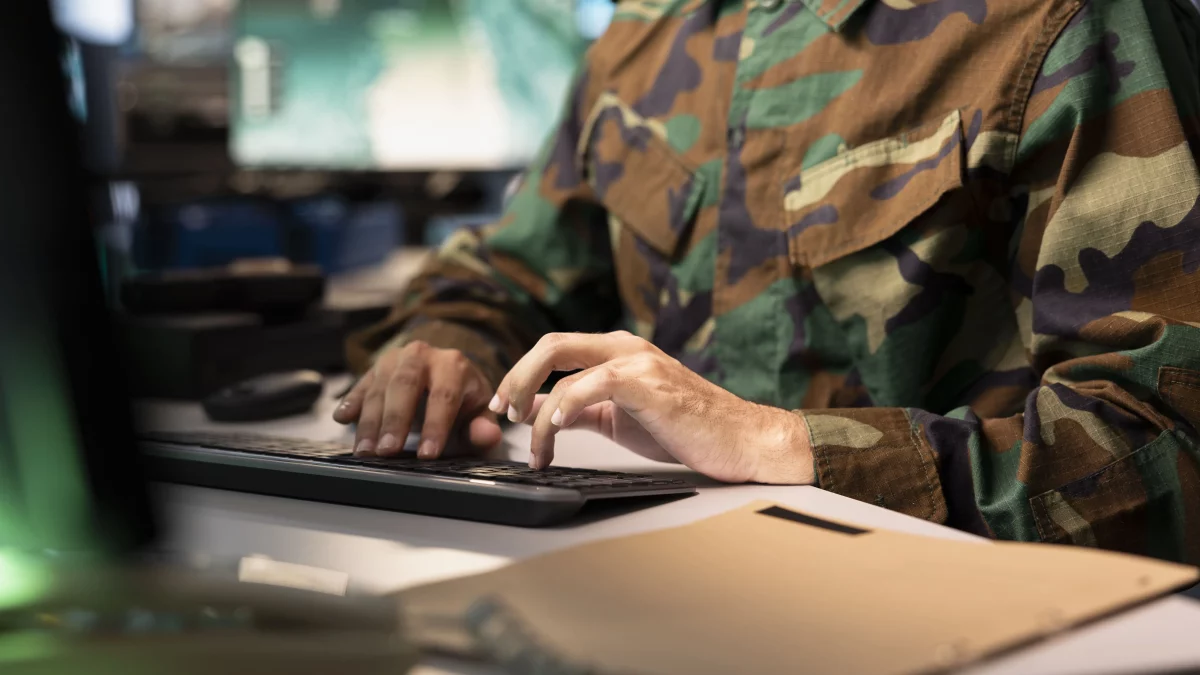 Militar de uniforme camuflado digitando em computador ao lado de documentos, representando erros no cálculo da pensão militar e possíveis cobranças administrativas.