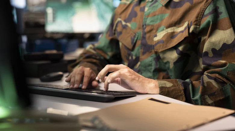 Militar de uniforme camuflado digitando em computador ao lado de documentos, representando erros no cálculo da pensão militar e possíveis cobranças administrativas.
