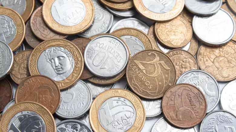 Fotografia em plano zenital (vista de cima) mostrando uma grande quantidade de moedas da moeda corrente brasileira, o Real, espalhadas e sobrepostas de forma desordenada. É possível identificar moedas de 1 Real com suas bordas douradas e centros prateados, moedas de 50 e 10 centavos prateadas, e moedas de 25 e 5 centavos em tons bronzeados. A imagem ilustra o conceito de frações de valores, acertos de contas e o impacto financeiro discutido no processo de devolução da pensão militar.
