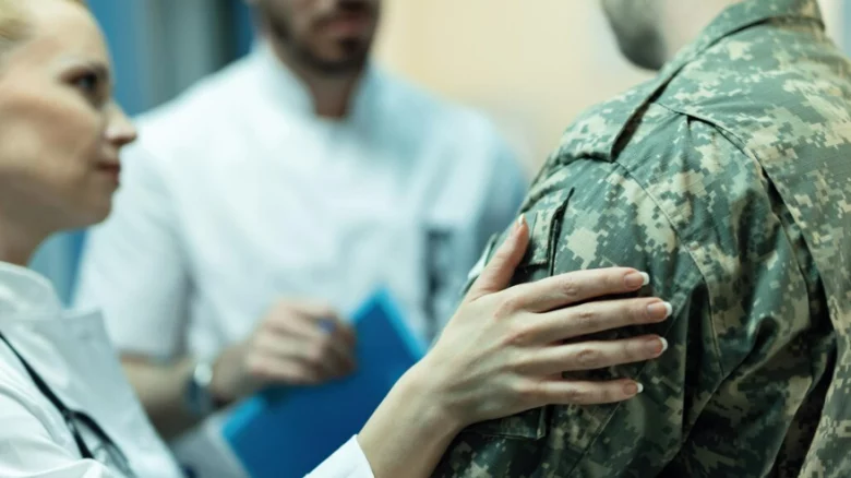Fotografia em plano médio focada nas costas e no ombro de um soldado usando farda camuflada digital em tons de verde. À sua frente, uma médica loira de jaleco branco apoia a mão em seu ombro em um gesto de apoio e acolhimento. Ao fundo, ligeiramente desfocado, um médico também de jaleco segura uma pasta azul. A cena ilustra o momento de uma consulta, inspeção de saúde ou acolhimento médico discutido no artigo sobre militares temporários em tratamento.
