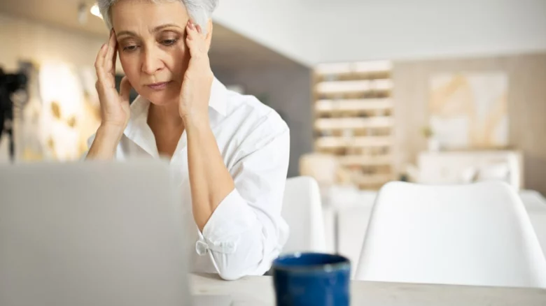 Uma mulher madura, de cabelos curtos e grisalhos, vestindo uma camisa branca, sentada à mesa em frente a um laptop. Ela está com uma expressão de profunda preocupação e cansaço, com os olhos fixos na tela e as mãos pressionando as têmporas, sugerindo dor de cabeça ou estresse ao ler uma notícia difícil. Em primeiro plano, o laptop e uma caneca azul aparecem ligeiramente desfocados, mantendo o foco no sentimento de angústia da mulher diante do computador.