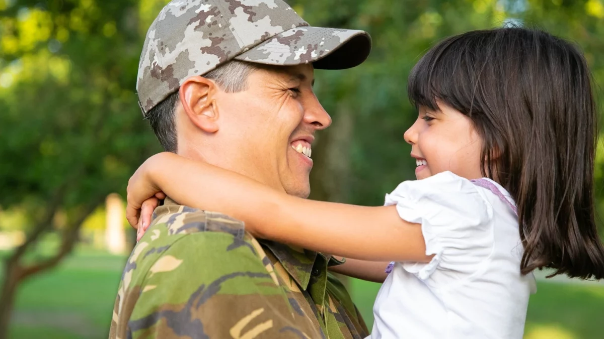 Um militar fardado, sorrindo e usando boné camuflado, segura nos braços sua filha pequena, que também sorri e o abraça pelo pescoço. O cenário é um ambiente externo arborizado, como um parque. A imagem ilustra o vínculo familiar e a preocupação do militar com o futuro de sua dependente, tema central para a discussão sobre pensão para filhas de militares.