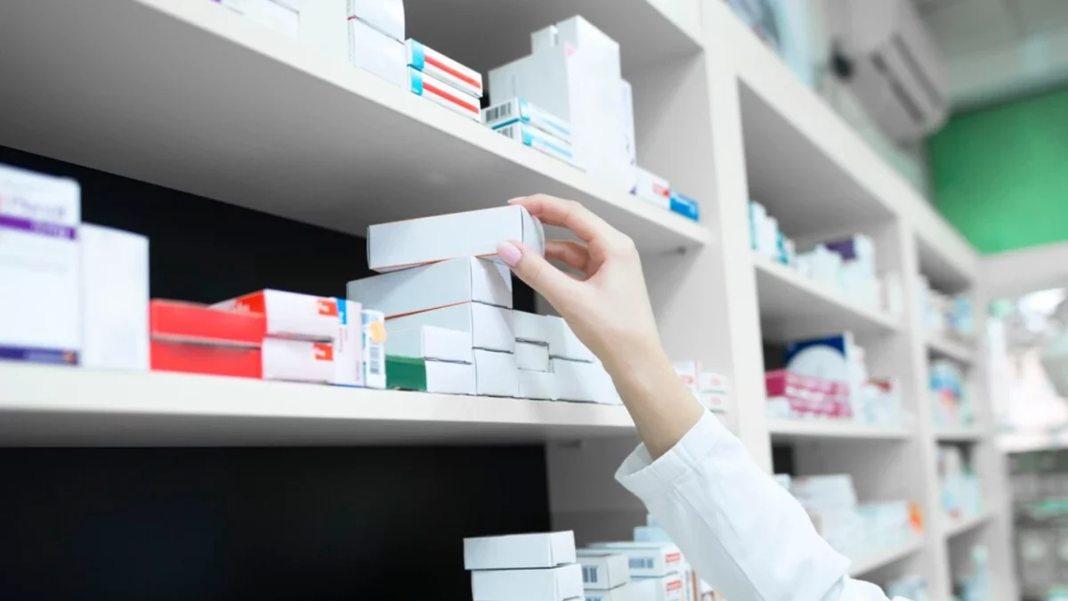 Visão em ângulo baixo em uma farmácia de prateleiras brancas repletas de caixas de medicamentos. A mão de um farmacêutico de jaleco branco pega uma caixa branca de uma pilha. Uma imagem que simboliza o acesso a medicamentos, relevante em discussões sobre negativas de planos de saúde para medicamentos de alto custo.