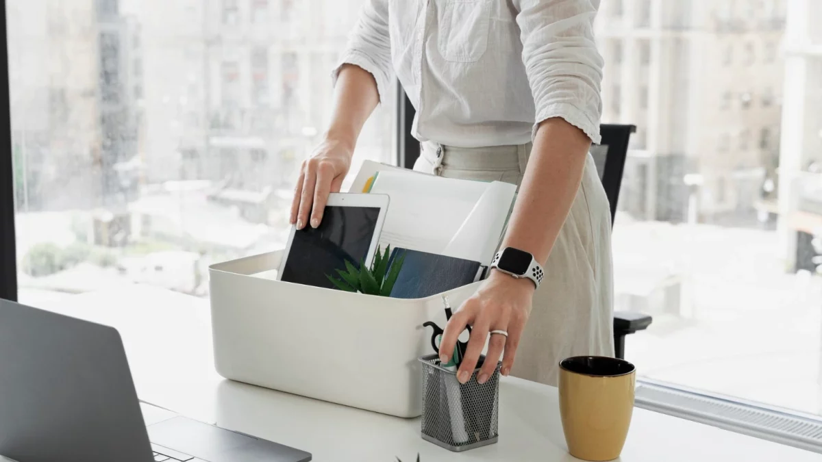 Uma profissional de camisa branca organiza seus pertences pessoais — como tablet, cadernos e porta-canetas — dentro de uma caixa branca sobre a mesa de escritório. Ao lado de um notebook aberto e de uma caneca, a cena retrata o momento de encerramento de um ciclo profissional, representando visualmente o processo de pedido de demissão ou rescisão indireta no ambiente de trabalho.