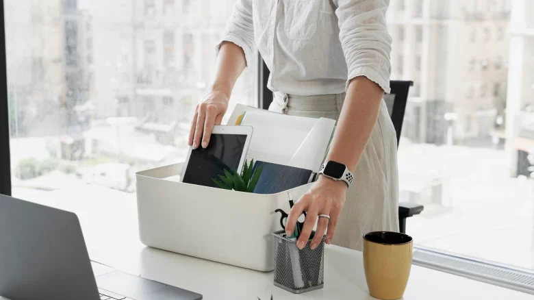Uma profissional de camisa branca organiza seus pertences pessoais — como tablet, cadernos e porta-canetas — dentro de uma caixa branca sobre a mesa de escritório. Ao lado de um notebook aberto e de uma caneca, a cena retrata o momento de encerramento de um ciclo profissional, representando visualmente o processo de pedido de demissão ou rescisão indireta no ambiente de trabalho.