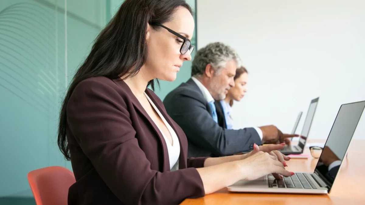 Uma mulher de óculos e blazer marrom está sentada em primeiro plano em uma mesa de escritório de madeira, digitando concentrada em um laptop aberto. Atrás dela, em uma fileira, estão um homem de terno cinza e outra mulher desfocada, também trabalhando em seus computadores. A cena reflete o ambiente de trabalho de servidores públicos em processos administrativos e a análise de dados que podem impactar a aposentadoria.