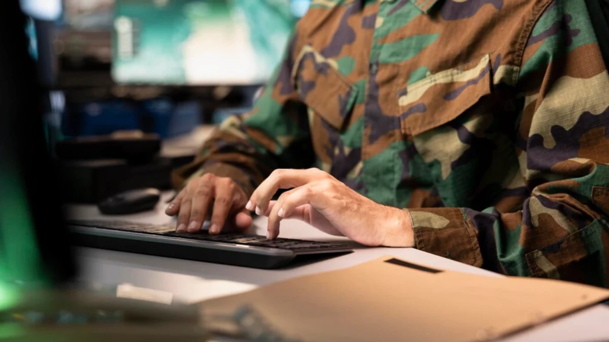 Uma imagem em plano médio foca nas mãos de um soldado vestindo uma jaqueta de camuflagem militar padrão, digitando em um teclado preto de computador sobre uma mesa branca em um ambiente de escritório. O soldado está sentado, mas sua cabeça e rosto não estão visíveis, apenas o tronco. No canto inferior esquerdo, há um monitor de computador preto e, à direita, uma pasta de papel pardo e um teclado.
