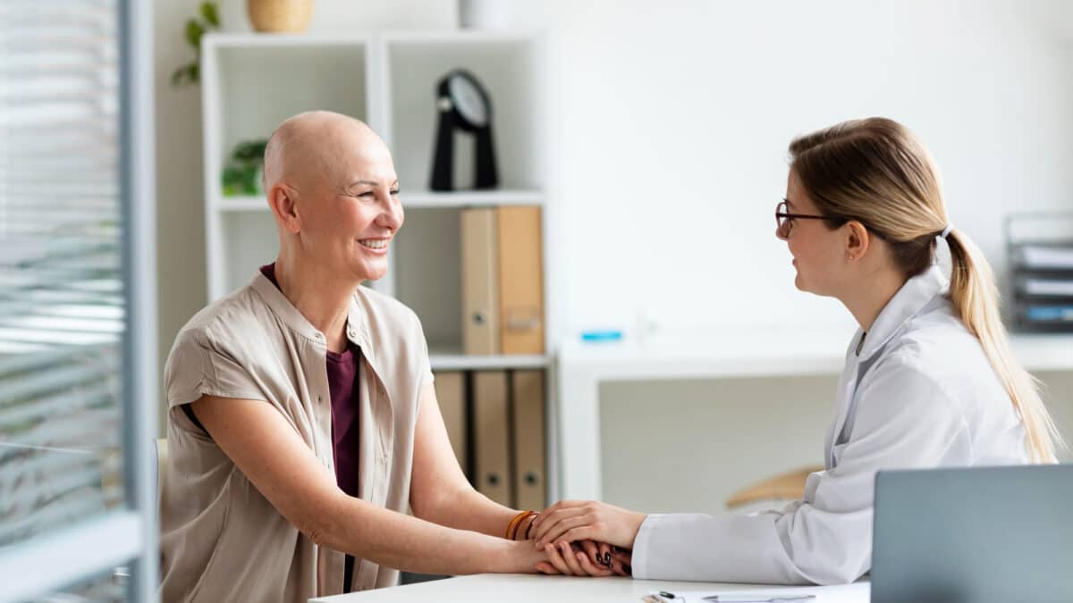 Imagem de uma mulher com câncer em uma consulta com uma médica, as duas conversando, sorrindo e de mãos dadas para simbolizar os direitos do paciente com câncer garantidos por lei