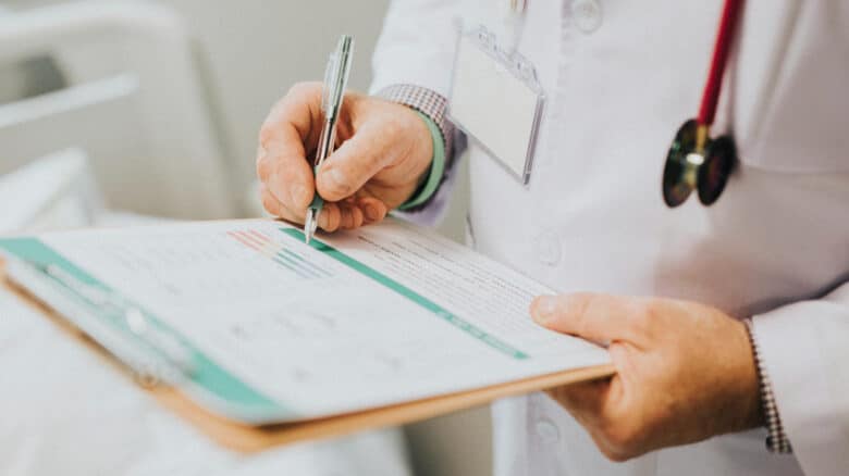 Imagem de um médico com umas folhas em uma prancheta nas mãos, anotando alguma coisa para simbolizar o dependente no plano de saúde